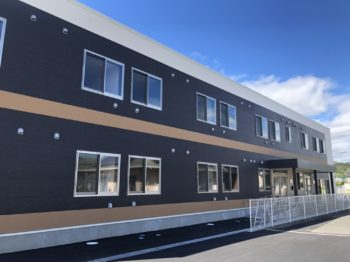 黒と茶色の外壁を持つ介護施設の建物外観。晴天下での全体像。